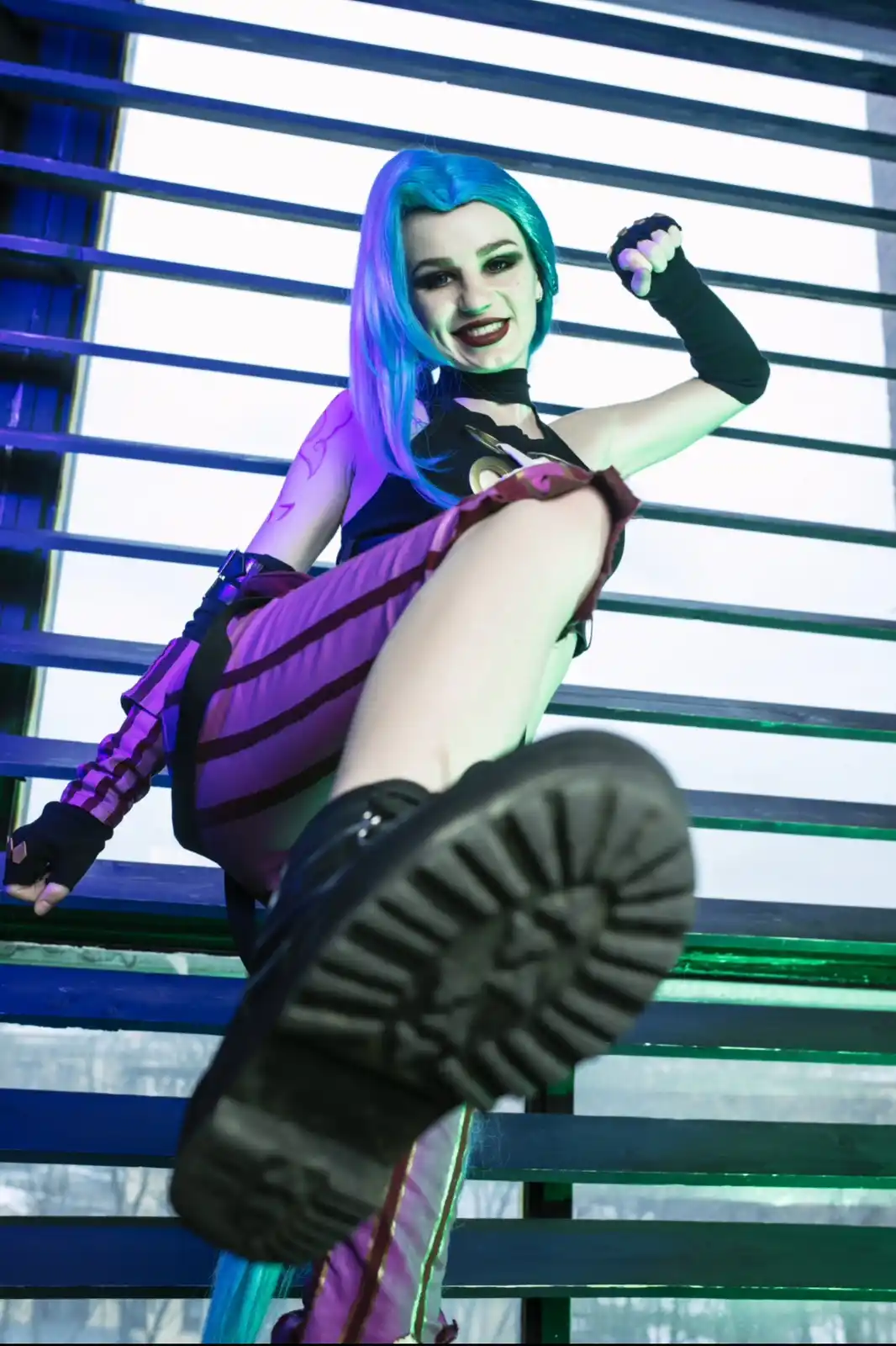 A person in a cosplay of the character Jinx from League of Legends poses confidently with one leg kicked up, showing the…