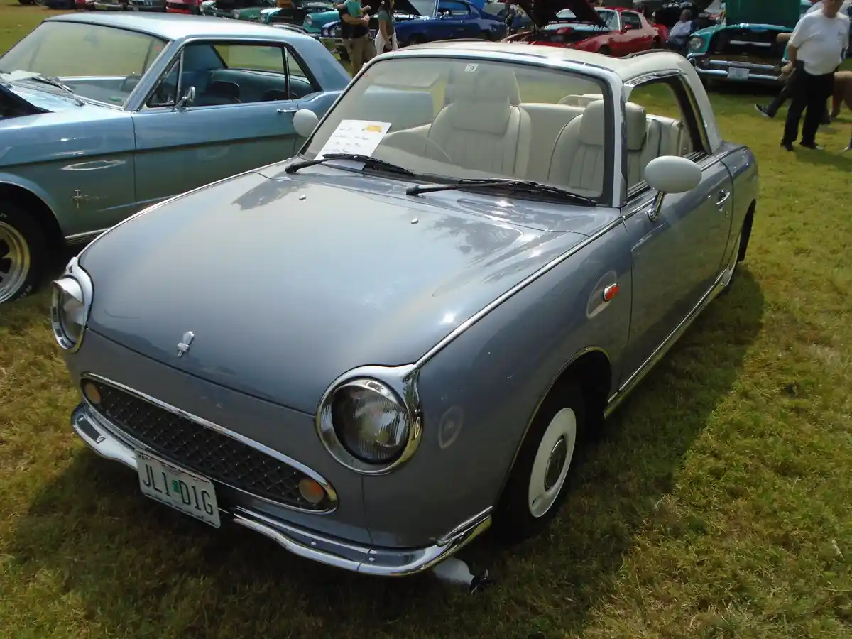 A light blue, retro-styled Japanese convertible car with white interior and chrome accents is parked on grass at a car show…