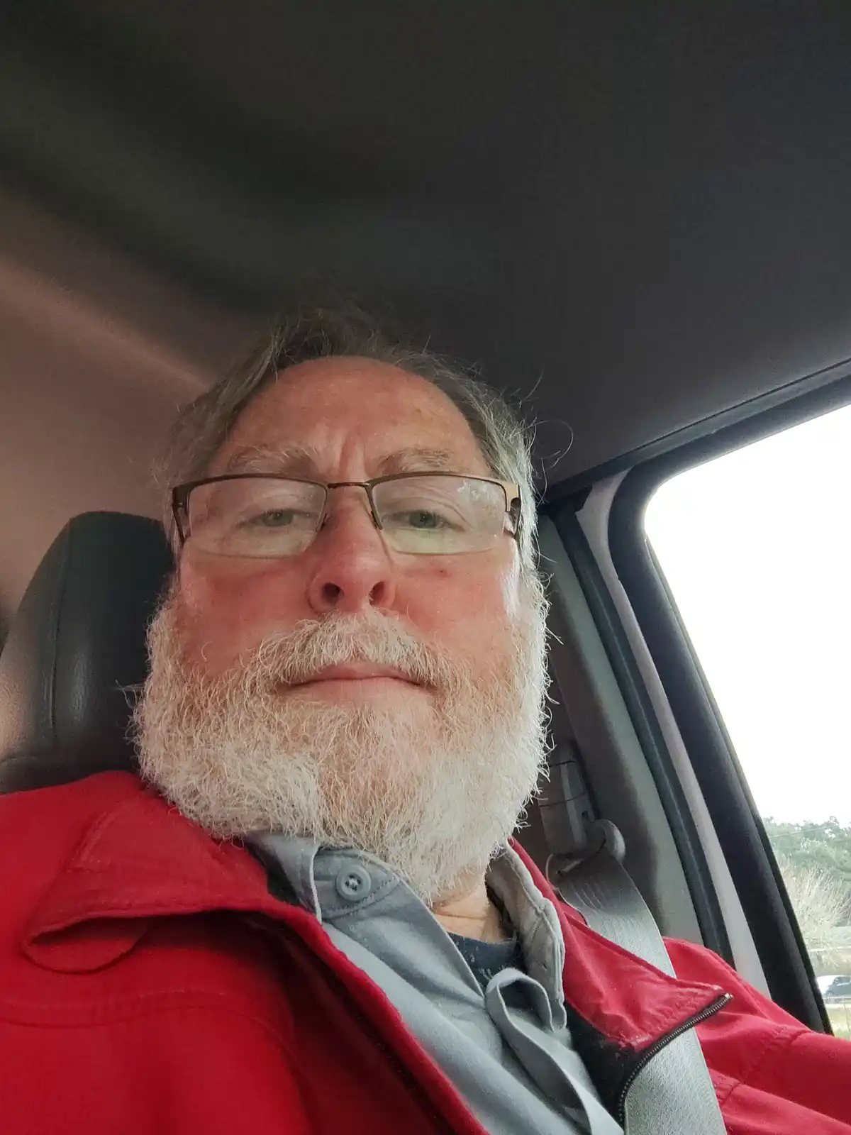 A man with a white beard and glasses sits in a car seat, wearing a red jacket over a grey shirt, looking directly at the…