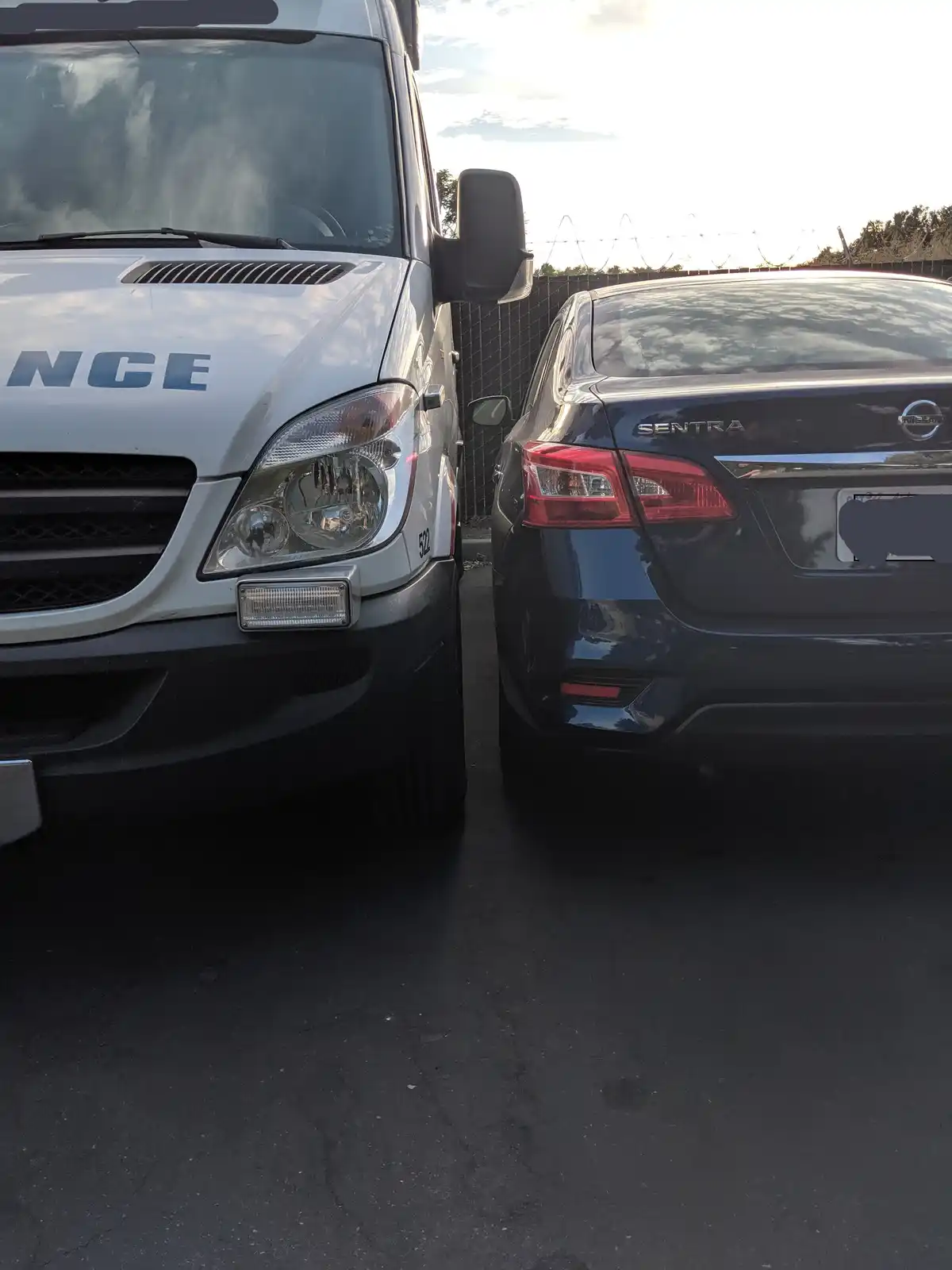 A white police van with 'NCE' visible on the hood is parked directly beside a dark blue Nissan Sentra, which has its rear…