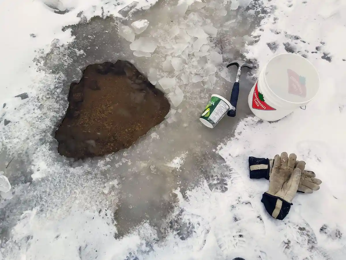 An overhead shot shows a hole in a frozen, snow-covered surface with muddy water, surrounded by broken ice. Nearby lie a…