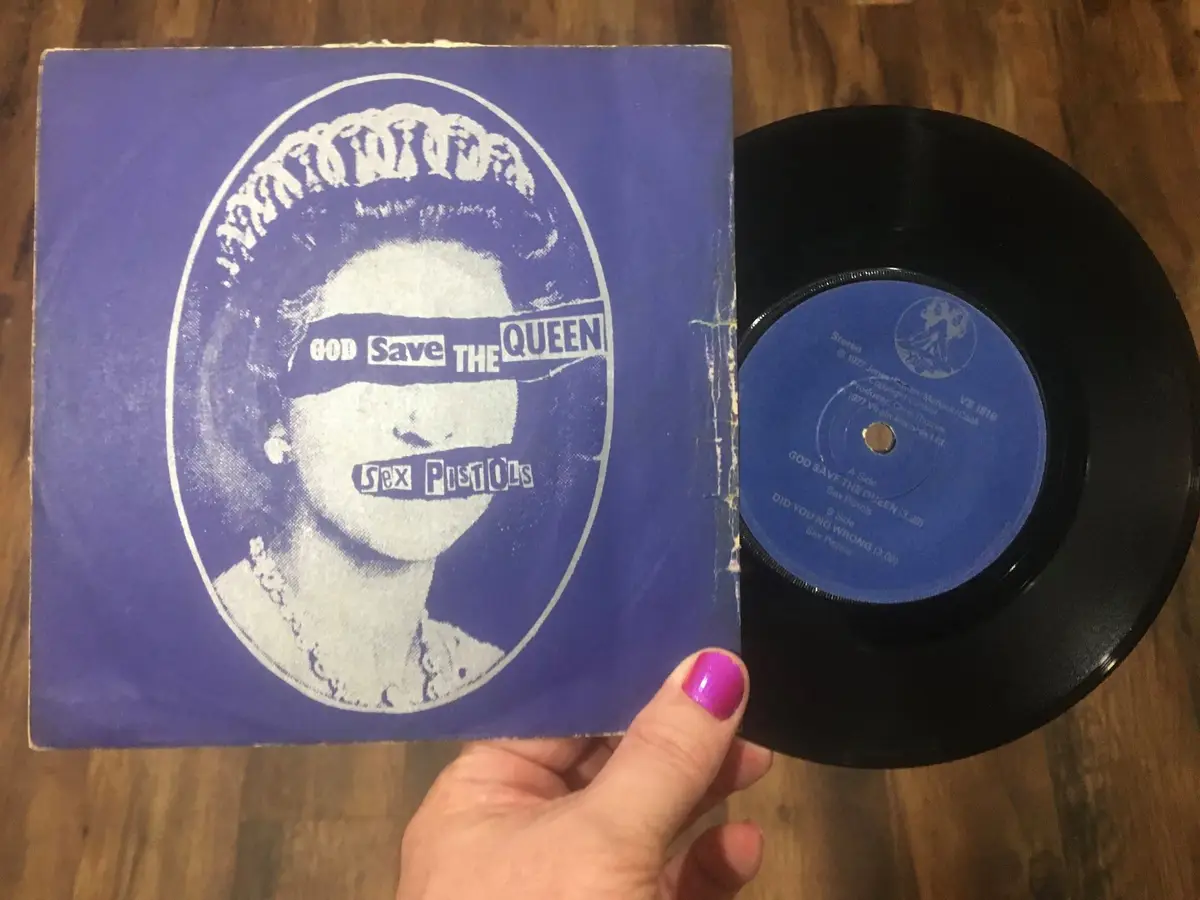 A hand holds a vintage Sex Pistols 7-inch vinyl record and its blue sleeve, which features a stylized portrait of the Queen…