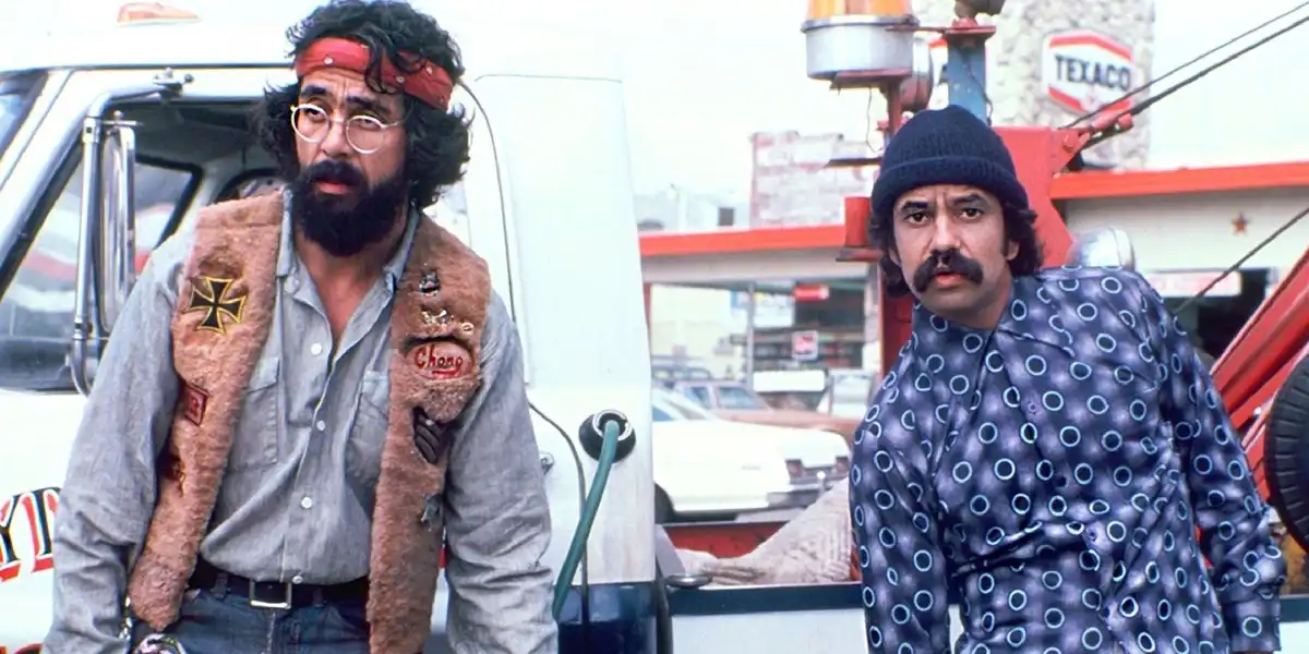 Two men from the movie 'The Big Lebowski' stand at a gas station; one wears a red bandana and glasses with a 'Chong' vest…