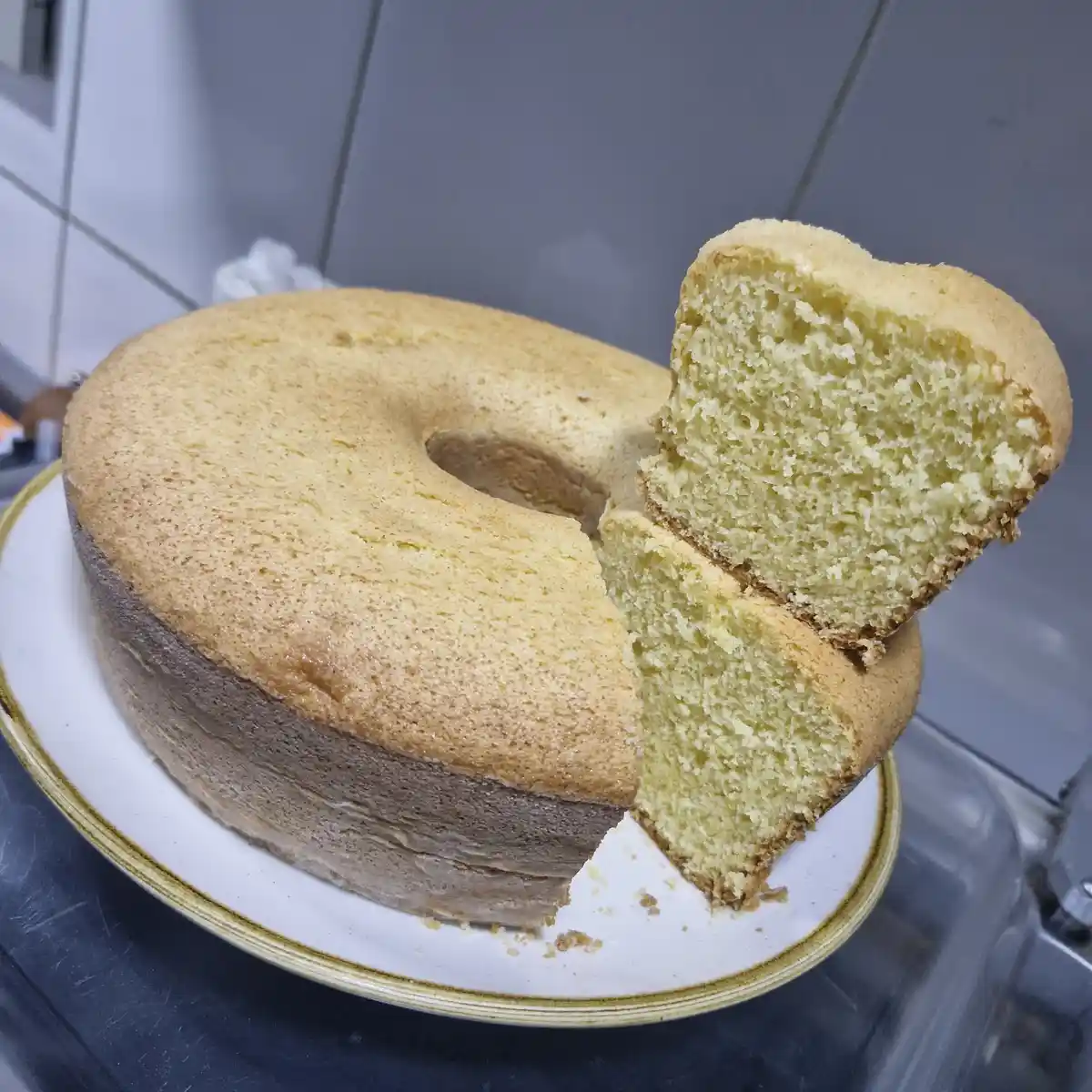 A golden-brown bundt cake sits on a white plate with a gold rim, one slice cut out and standing upright, revealing a pale…
