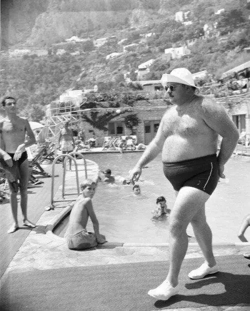 A black-and-white photo shows a large man in swim trunks and a white hat walking near a pool, with other people swimming and…