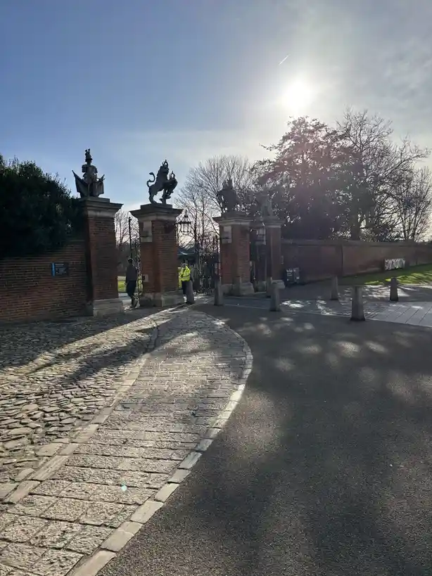A photo of a grand, ornate brick gate with statues on top, under a bright sun, with a cobblestone path leading up to it. A…