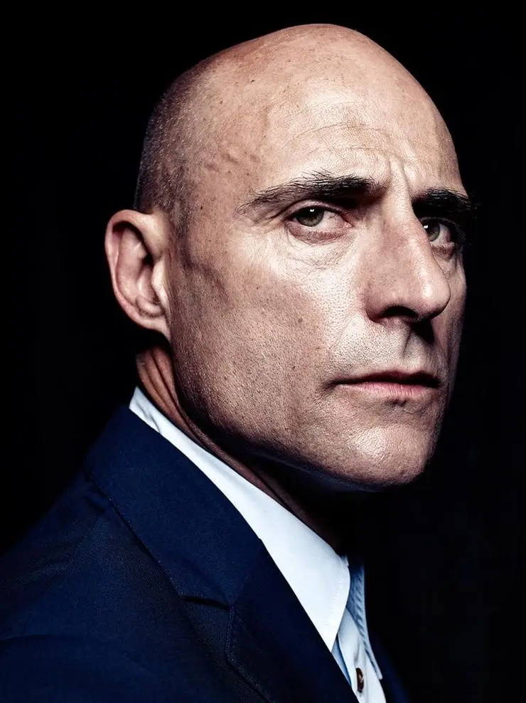 a close-up portrait of a bald man with intense eyes, wearing a dark suit and white shirt against a black background. The…