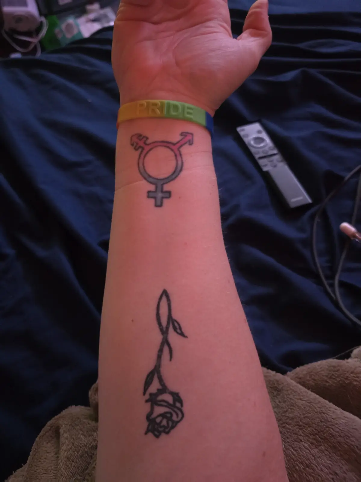 A person's forearm displays two tattoos: a genderqueer symbol near the wrist and a black rose tattoo lower down. A…