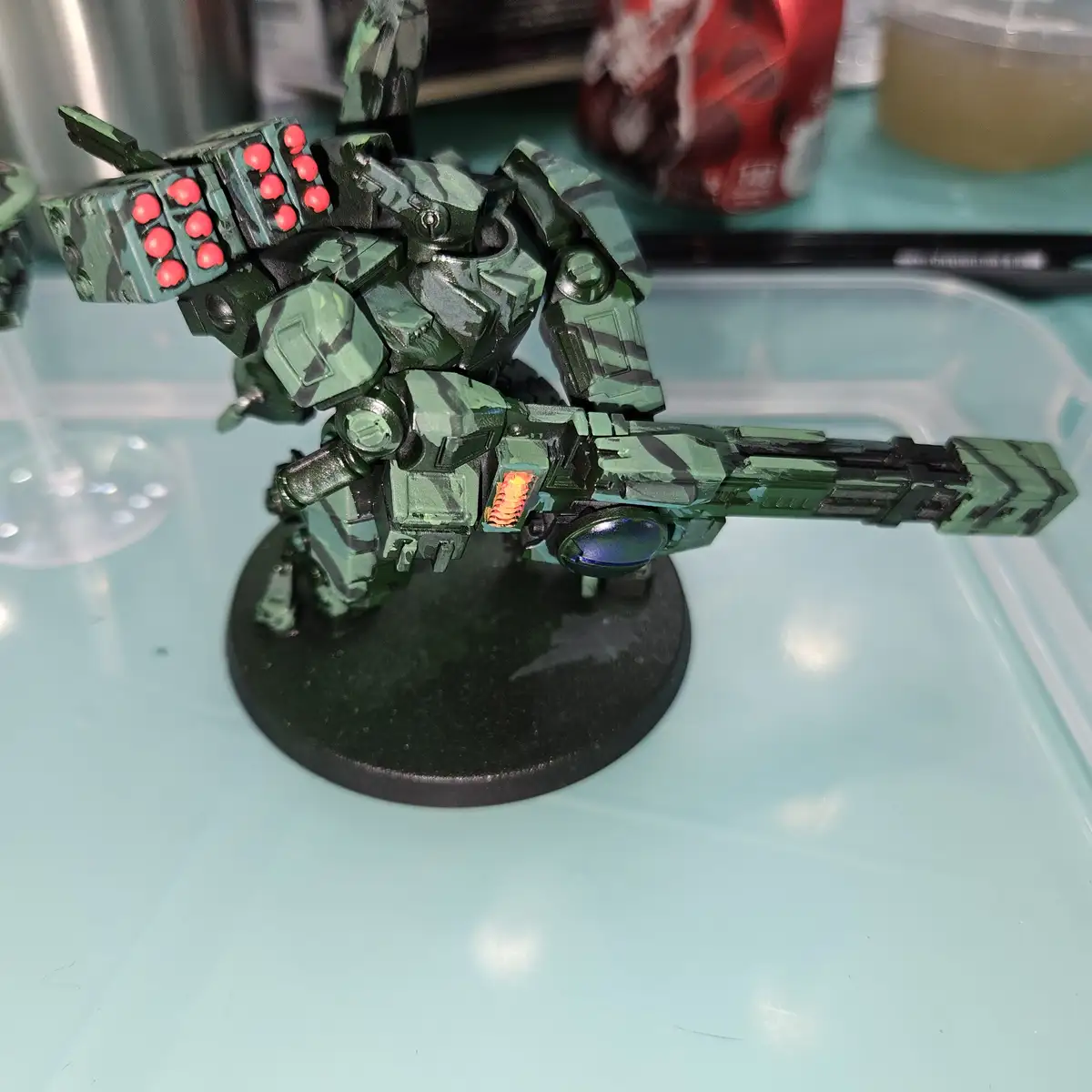 A green and black camouflaged robot model with red glowing orbs on its shoulder and a glowing orange chest panel stands on a…