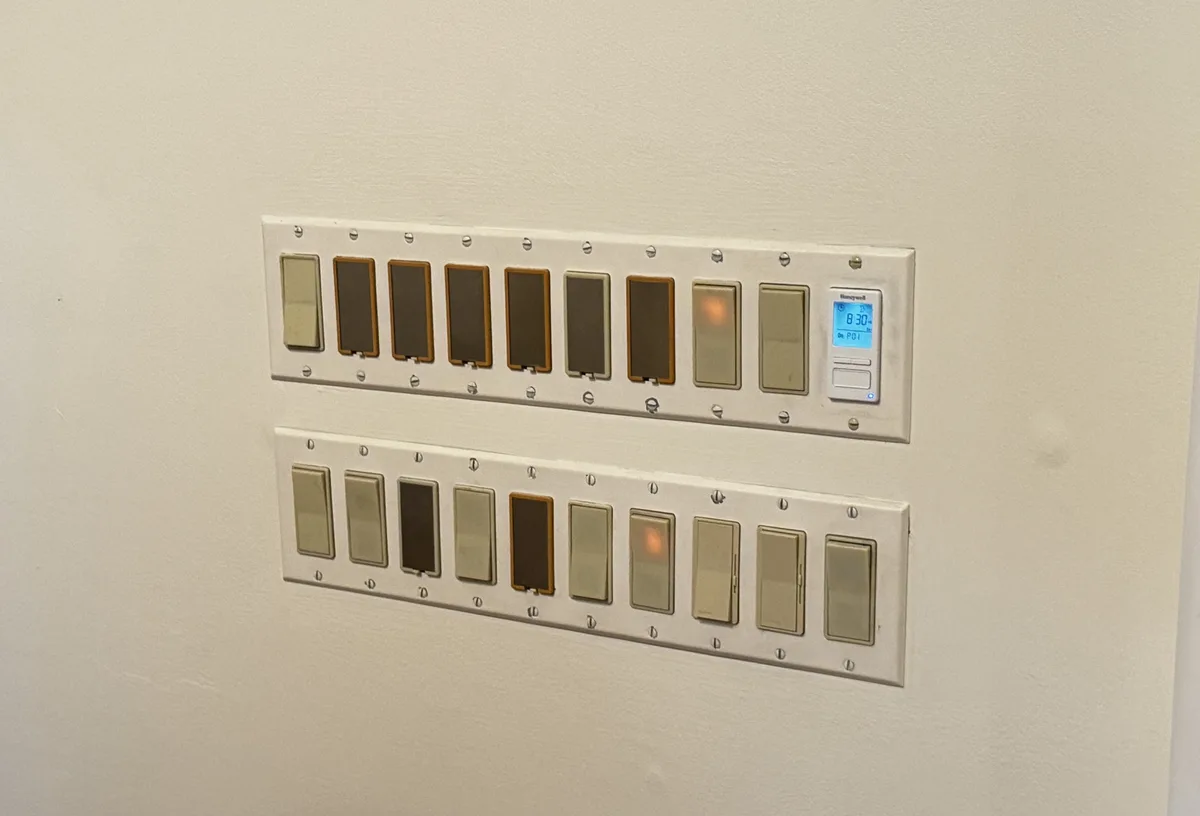 A wall features two rows of light switches, each with multiple dimmer switches and one digital thermostat displaying '8:30'…