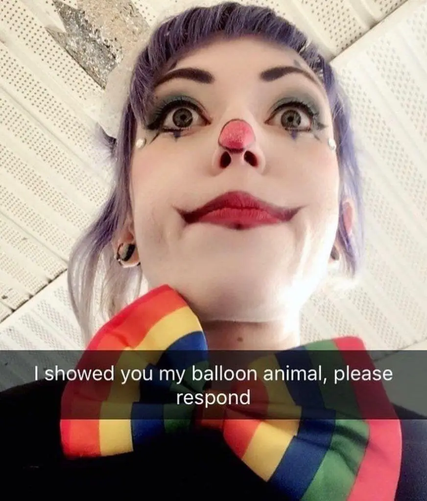 A person with purple hair, clown makeup including a red nose and white face paint, and a rainbow-striped bow tie looks…