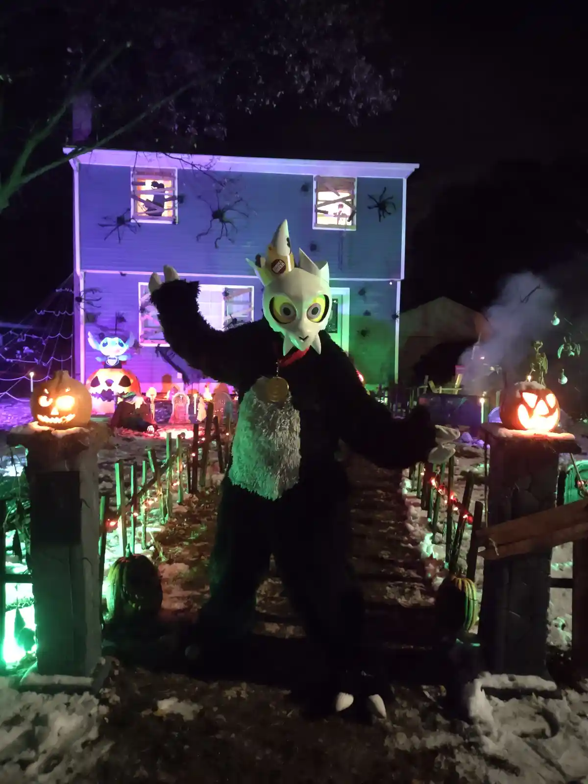A person in a black furry costume with a white owl-like mask and a crown stands in front of a house decorated for Halloween…