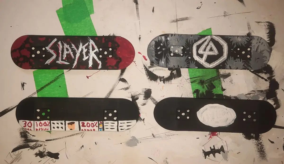Four hand-painted skateboard decks are arranged on a messy, graffiti-covered surface. The top-left deck has 'SLAYER' in…