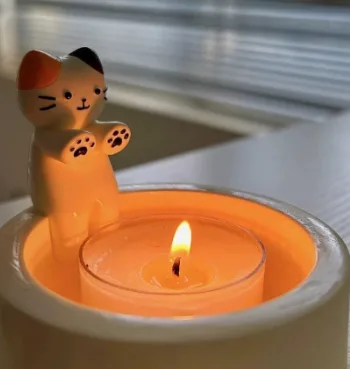 A small, white ceramic cat figurine with orange ears and black facial markings stands upright beside a lit, orange-colored…