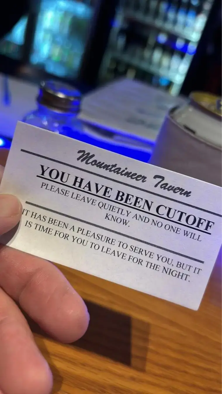 A hand holds a white card from 'Mountaineer Tavern' that reads 'YOU HAVE BEEN CUTOFF PLEASE LEAVE QUIETLY AND NO ONE WILL…