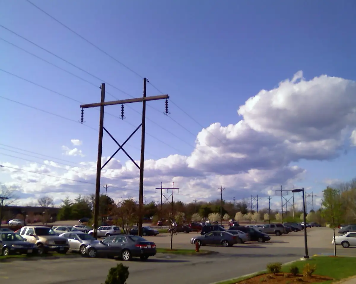 A daytime photo of a parking lot with cars, trees, and multiple wooden utility poles with power lines stretching across a…