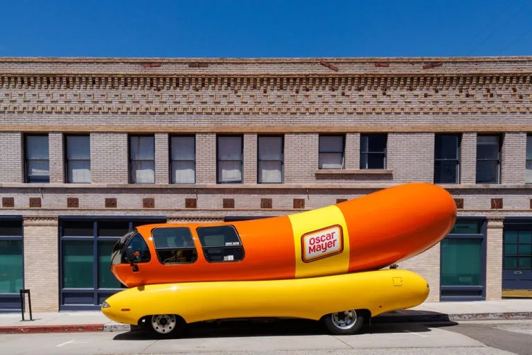 A large, iconic Oscar Mayer Wienermobile, shaped like a hot dog in a bun, is parked on a city street in front of a brick…