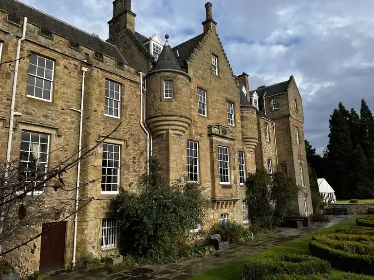 A large, historic stone castle with multiple turrets, windows, and a slate roof under a cloudy sky, with manicured gardens…