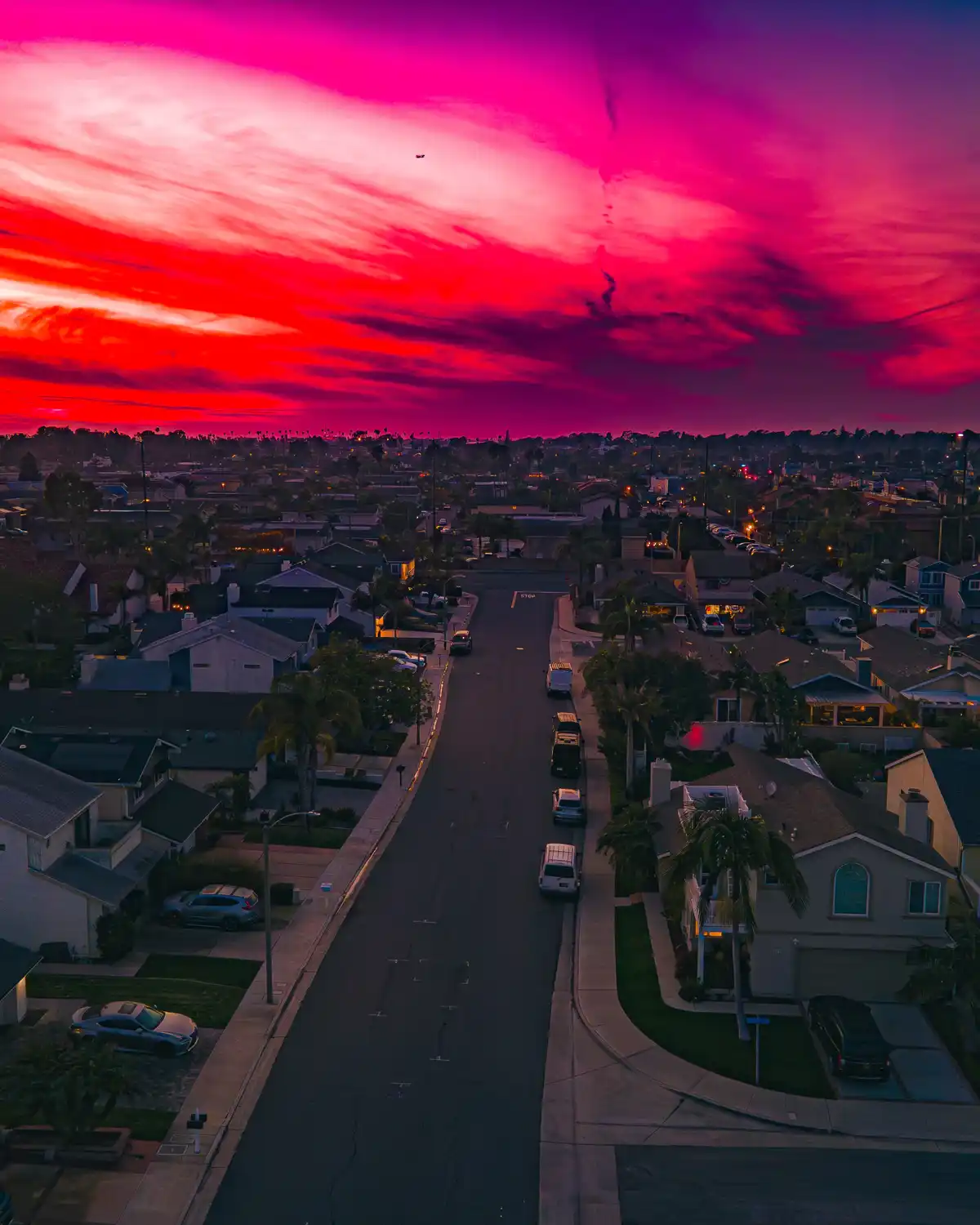 An aerial photo of a suburban street at dusk, with houses, parked cars, and palm trees under a sky saturated with vibrant…