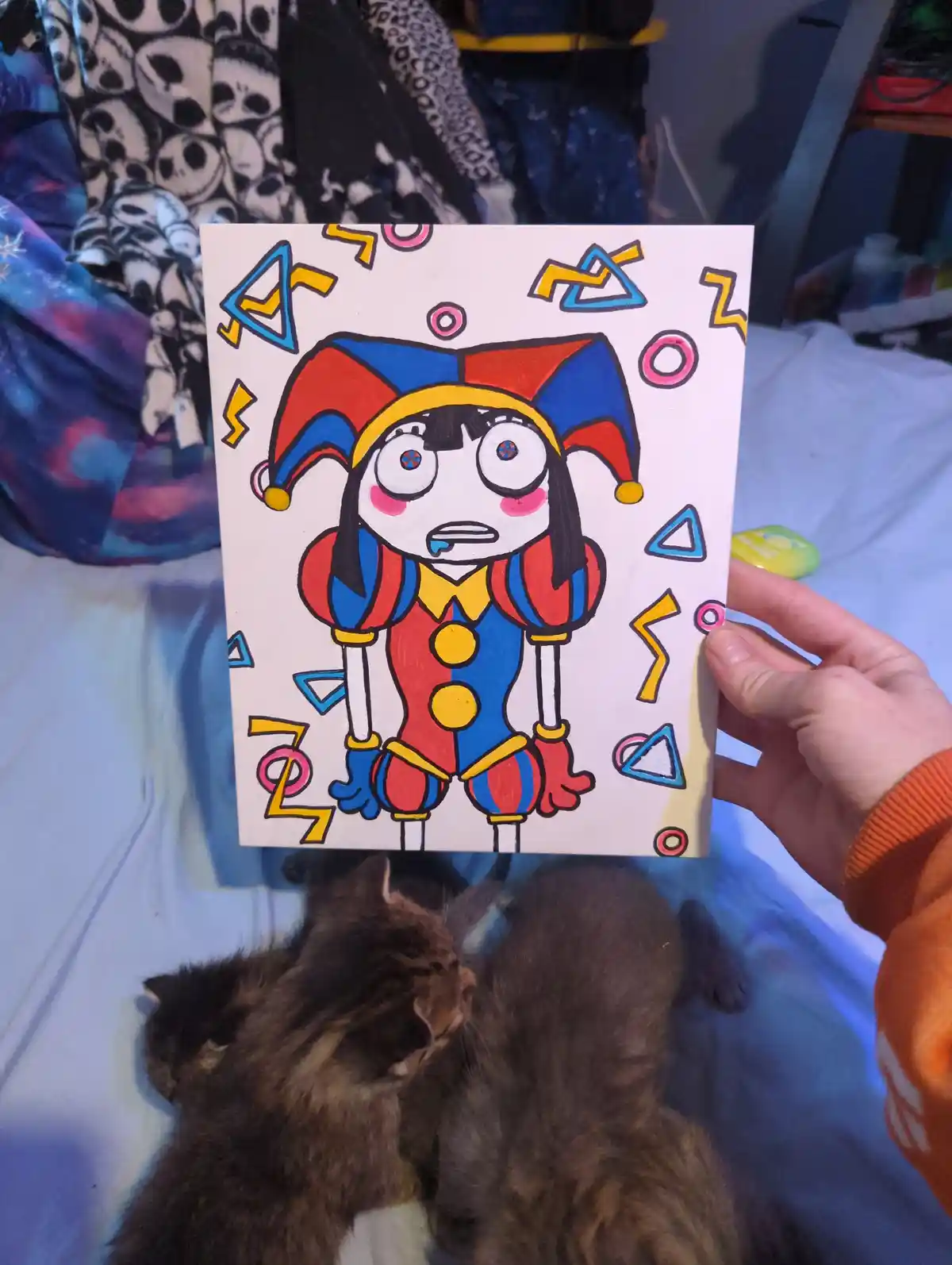 A hand holds up a colorful drawing of a cartoon clown with a red and blue hat, wide eyes, and a surprised expression…