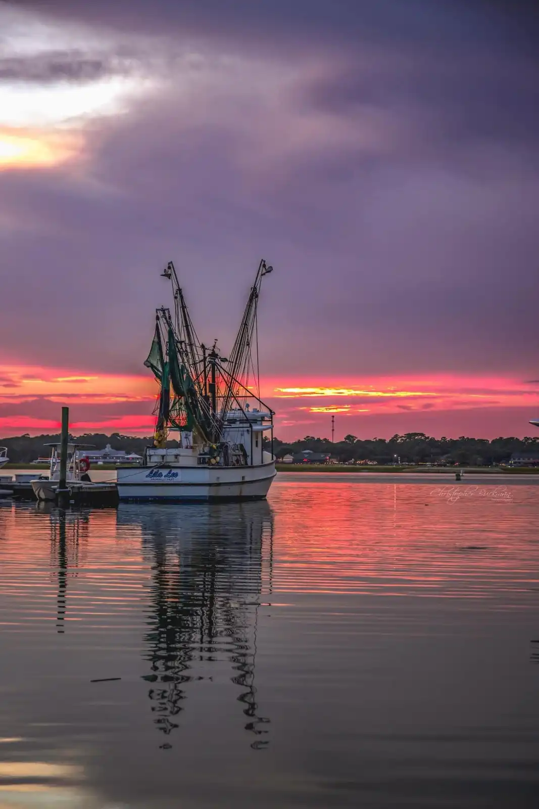 A serene photograph of a shrimp boat named 'Lila Lee' docked in calm water during a vibrant sunset, with purple and orange…