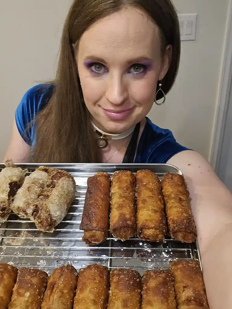 A woman with purple eyeshadow and a blue top holds a metal tray filled with golden-brown fried spring rolls, some of which…