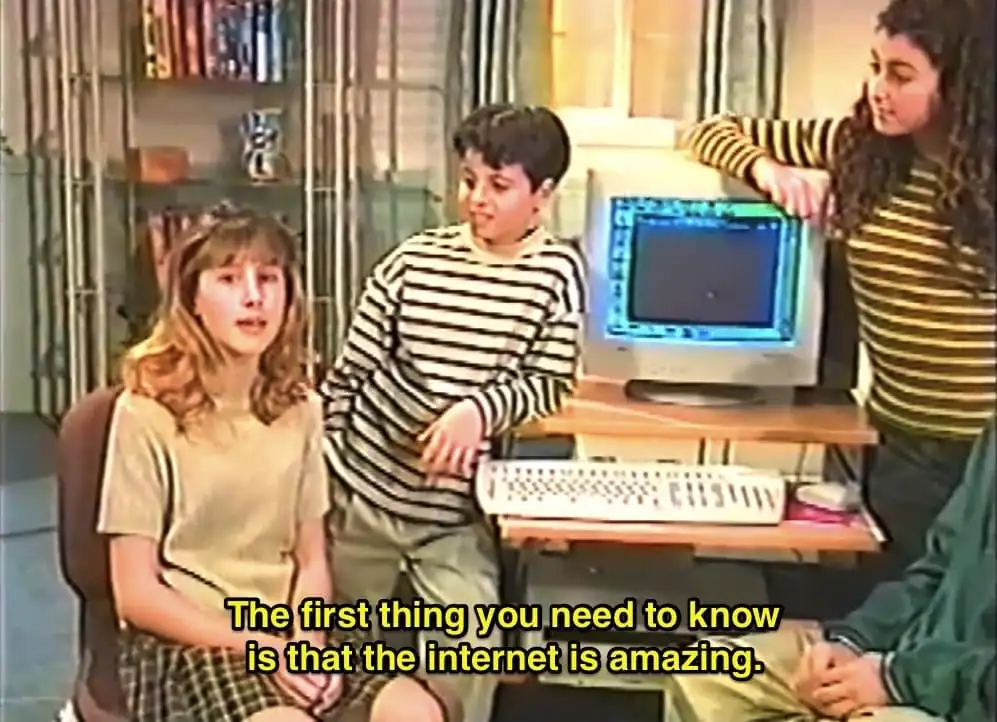 Three children in a 90s-style living room gather around an old beige computer with a blank screen. The girl on the left…