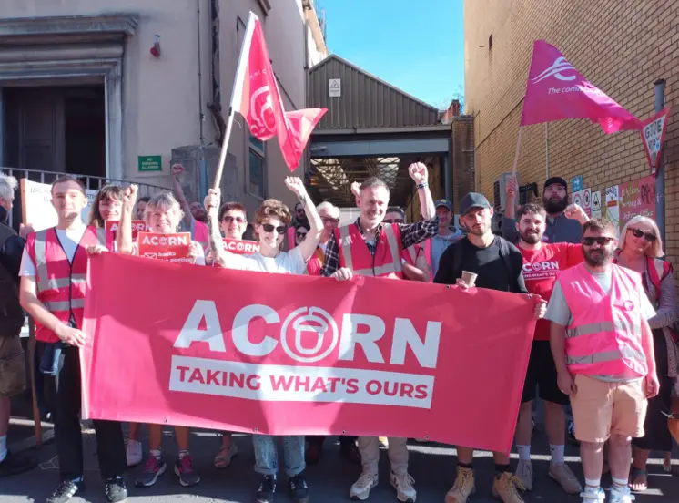A group of people wearing red and pink safety vests are protesting outdoors, holding a large red banner that reads 'ACORN…