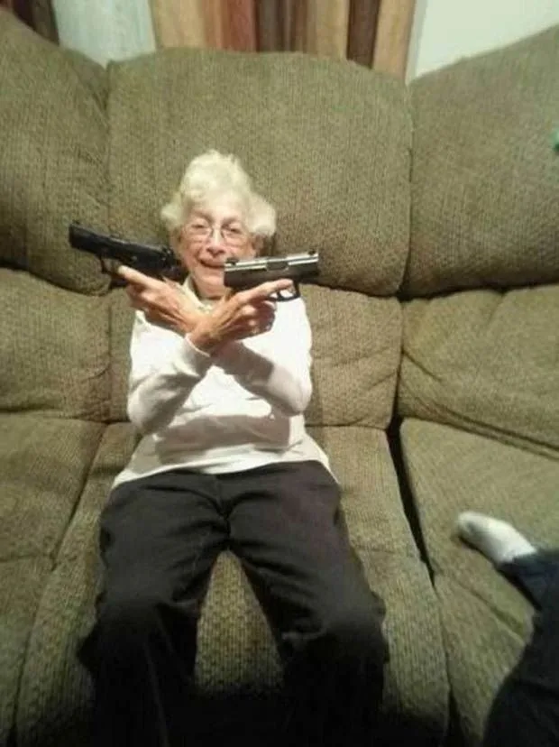 An elderly woman with white hair and glasses sits on a brown couch, holding two handguns, one in each hand, pointed outward…