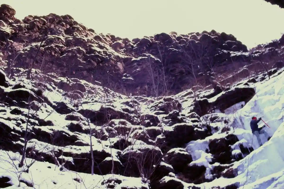 A person in winter gear is ice climbing on a snowy, rocky slope. The image has a vintage, slightly faded color tone…