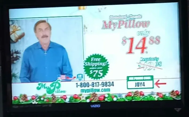 A television screen displays a MyPillow infomercial featuring a man in a blue shirt. The ad shows the pillow priced at…