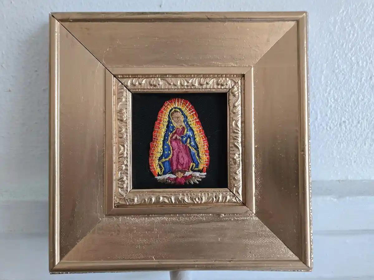 A small, hand-embroidered religious image of the Virgin of Guadalupe is displayed in a square, gold-painted wooden frame…