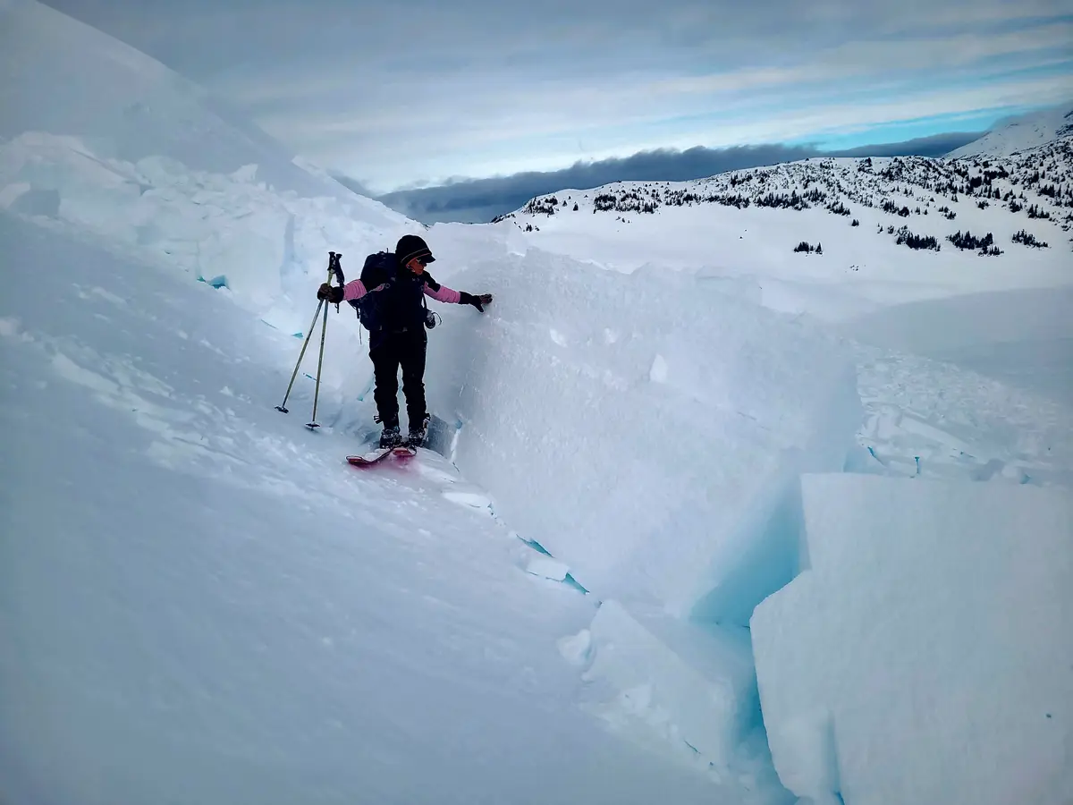 A skier in winter gear stands on a snowy slope next to a large, jagged snow wall, holding poles and touching the snow with…