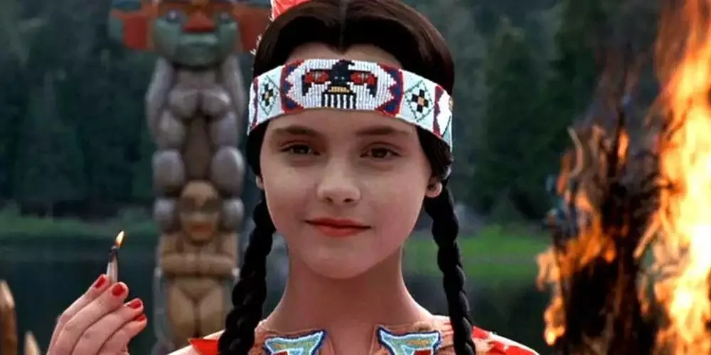 A young girl with braided hair and a beaded headband holds a lit match, standing before a fire and a totem pole. The…