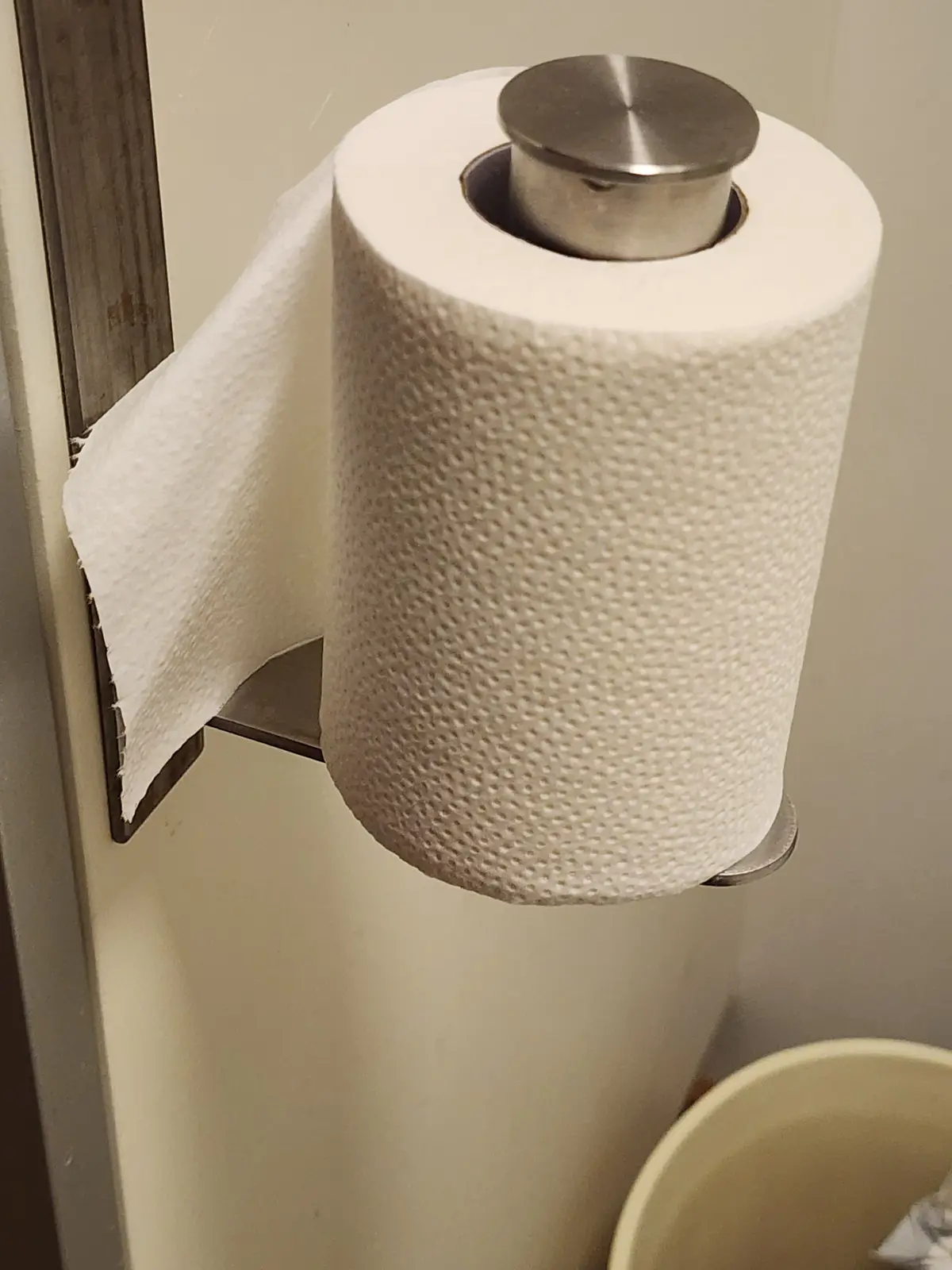 A close-up photo shows a large roll of textured white toilet paper on a modern metallic holder, with a single sheet pulled…
