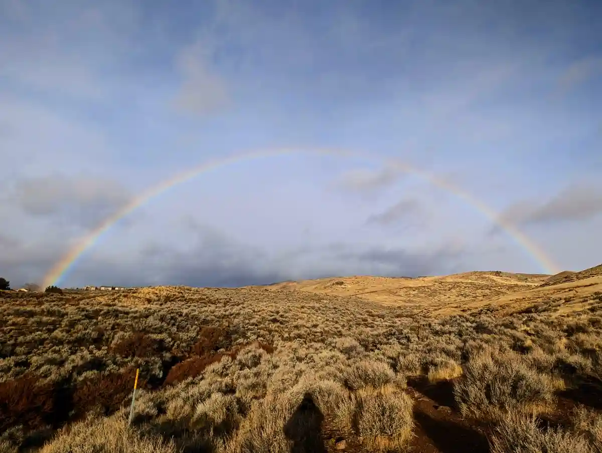 A wide-angle photo shows a vivid, full rainbow arcing across a partly cloudy sky above a dry, rolling landscape covered in…
