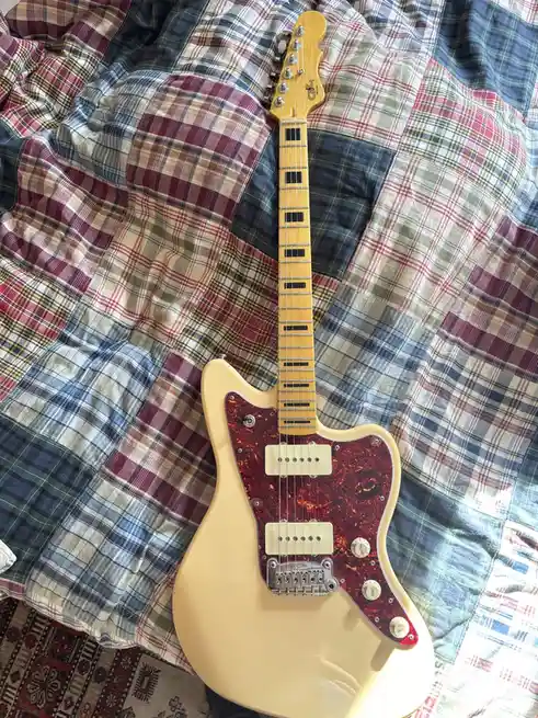 A light cream-colored electric guitar with a red tortoiseshell pickguard rests on a colorful patchwork plaid blanket. The…