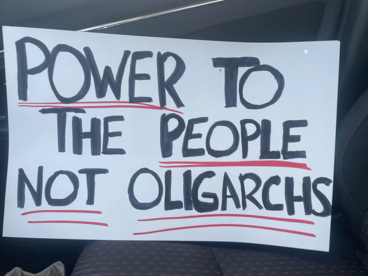 A white protest sign with bold black handwritten text reads 'POWER TO THE PEOPLE NOT OLIGARCHS', with each line underlined…