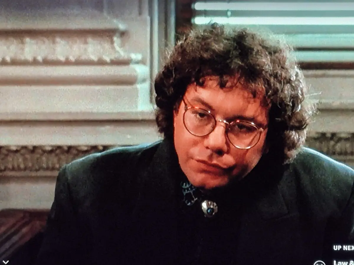 A still image of a man with curly hair and round glasses, wearing a dark coat with a decorative brooch, looking down with a…