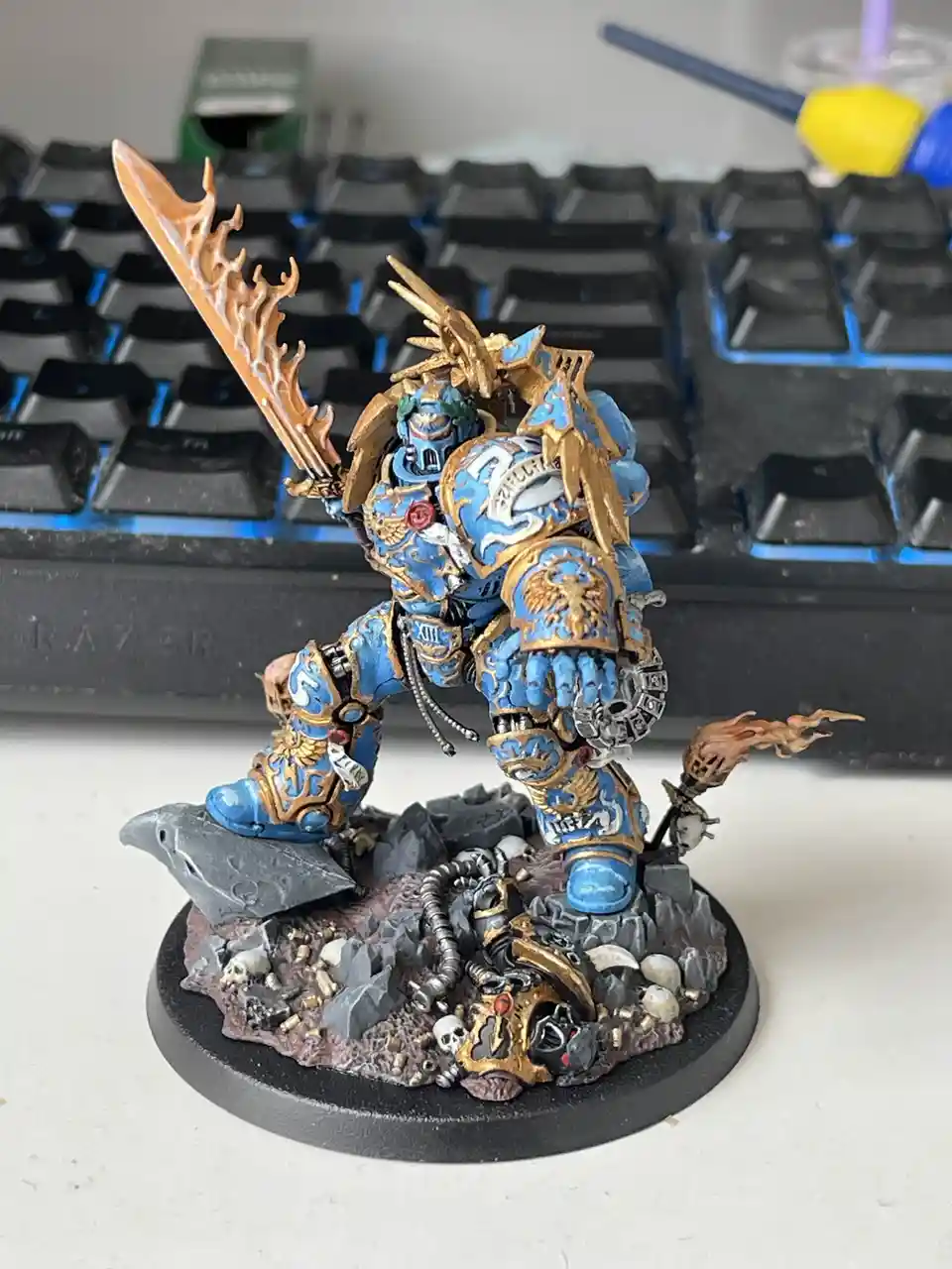 A highly detailed, blue-and-gold painted miniature of a Space Marine from Warhammer 40,000 stands on a rocky, skull-strewn…