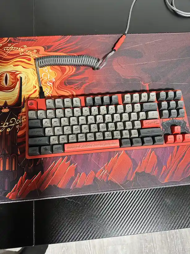 A mechanical keyboard with keys labeled in Elvish script sits on a matching Elvish-themed mousepad depicting fiery…