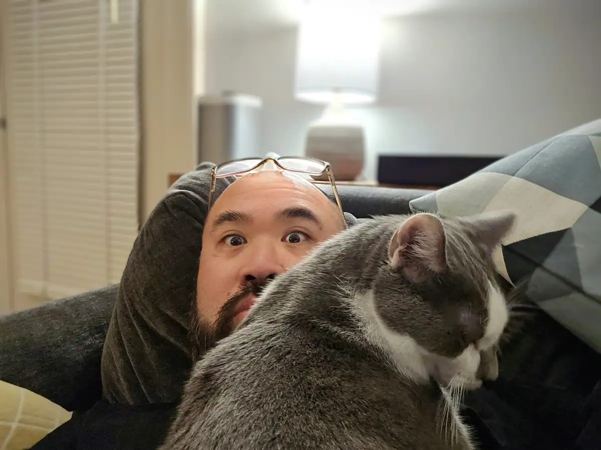 A man with glasses on his forehead lies on a couch, looking startled with wide eyes as a gray and white cat rests its head…