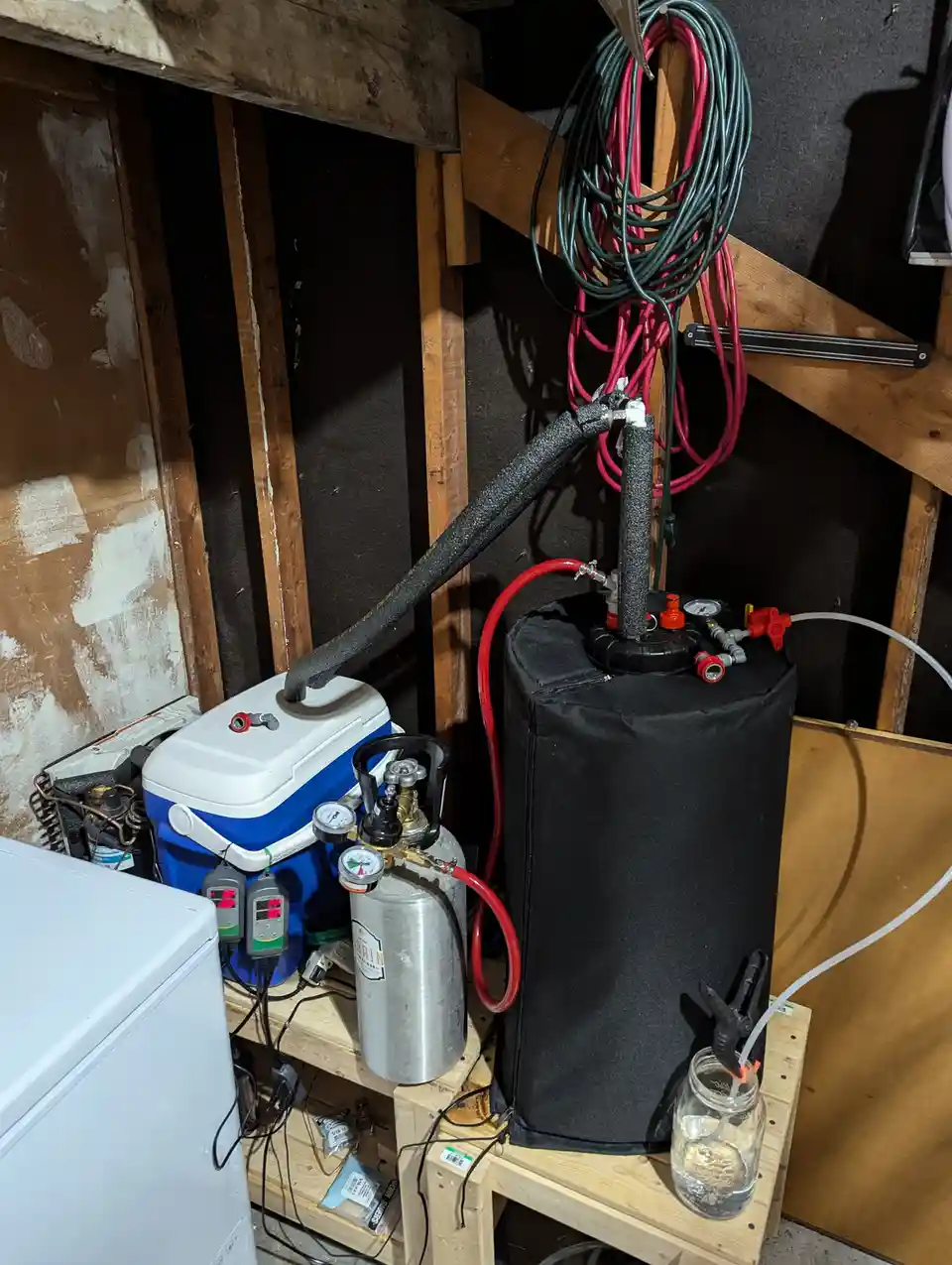 A cluttered basement setup features a blue and white cooler with gauges and a silver propane tank, connected by red hoses to…
