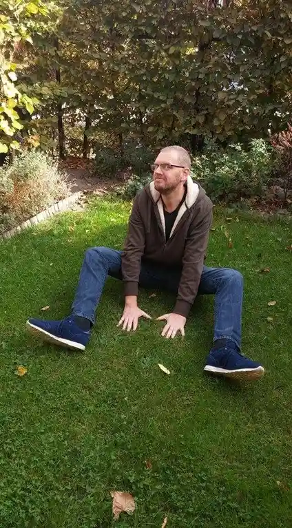 A man with glasses and a beard sits cross-legged on green grass in a garden, wearing a brown hoodie, jeans, and blue…