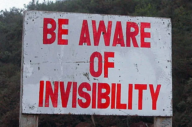 A weathered white sign with bold red text reads 'BE AWARE OF INVISIBILITY' against a backdrop of trees and foliage. The sign…