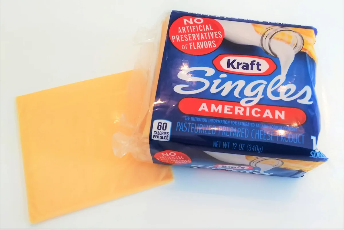 A package of Kraft Singles American cheese sits next to a single slice, with the text 'NO ARTIFICIAL PRESERVATIVES OR…