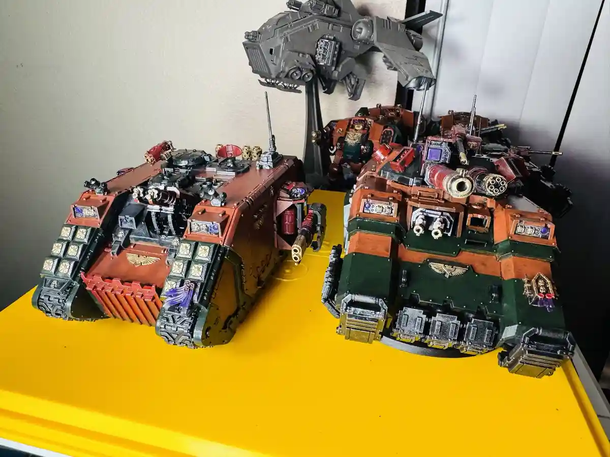Two highly detailed, custom-painted Warhammer 40,000 tank models sit side-by-side on a bright yellow surface, with a larger…