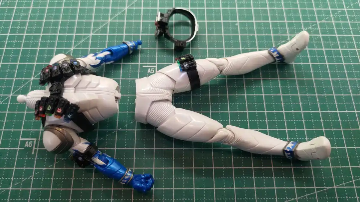 A disassembled white and blue action figure lies on a green cutting mat, with its limbs and accessories scattered around…