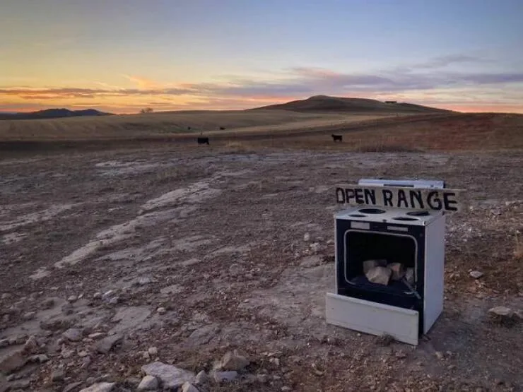A vintage white stove sits alone in a vast, rocky desert landscape at sunset, with distant black cattle grazing on the…