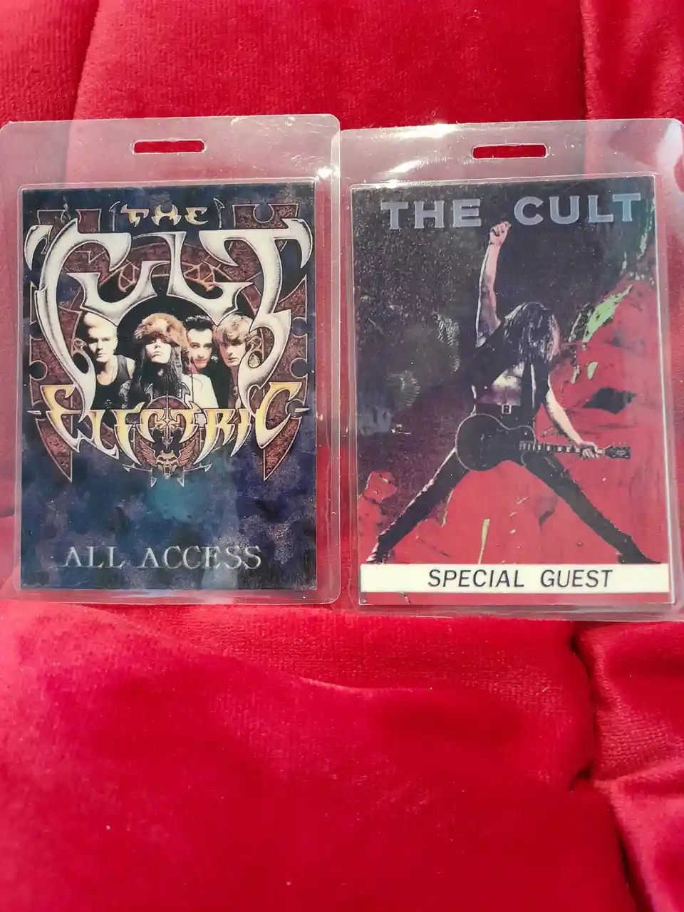 Two plastic badges lie side-by-side on a red fabric surface. The left badge features the band 'The Cult Electric' with 'ALL…
